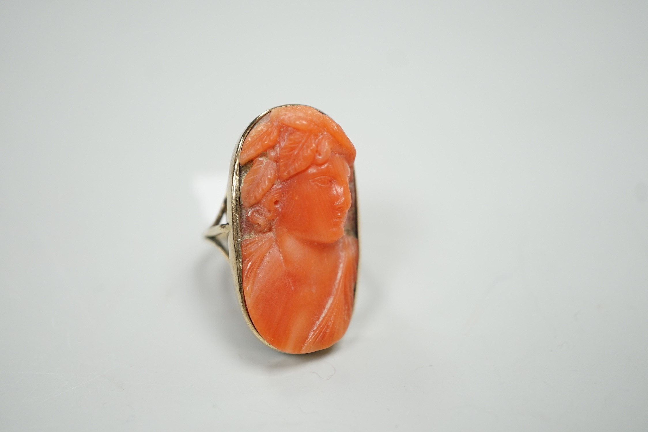 A yellow metal and coral set oval dress upfinger ring, the coral carved with a classical bust to sinister, size O, gross weight 8.2 grams.